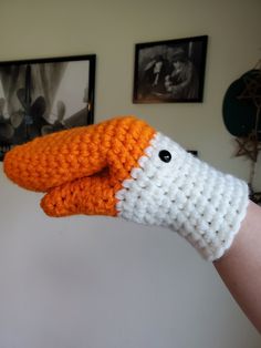 an orange and white knitted animal is held in someone's hand with two pictures on the wall behind it