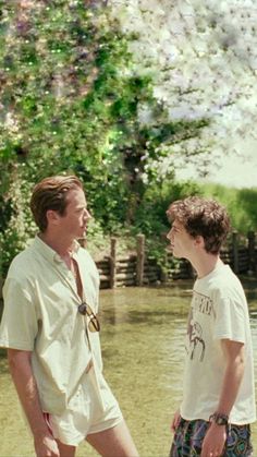 two men standing next to each other in front of a body of water with trees