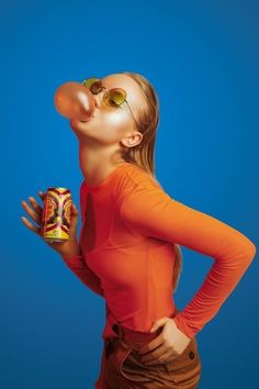 a woman in an orange shirt and sunglasses is holding a canned drink up to her mouth