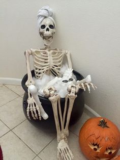 a skeleton sitting in a black bowl next to two pumpkins