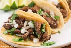 three tacos with meat, onions and cilantro on a white plate next to lime wedges