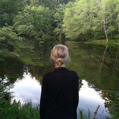 a woman standing in front of a body of water surrounded by green trees and bushes