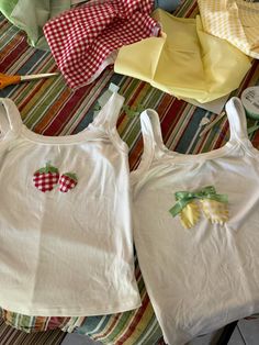 two white shirts with fruit appliques on them sitting on a striped table cloth