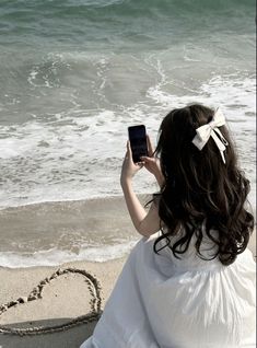 a woman in white dress sitting on the beach using her cell phone to take a photo