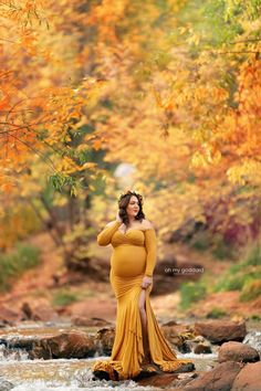 a pregnant woman in a yellow dress standing on rocks near a river with fall foliage