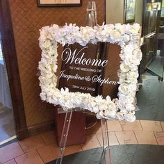 a welcome sign with white flowers on it in front of a door way at a wedding