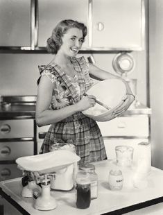 an old photo of a woman in the kitchen holding a bowl and mixing spoons