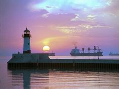 the sun is setting behind a lighthouse and cargo ship in the ocean with ships on the horizon