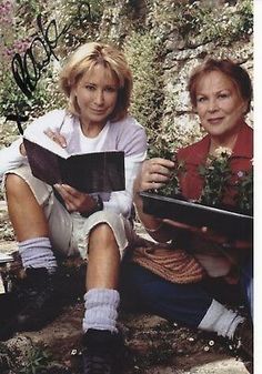 two women are sitting on the ground and one is holding an open book while the other holds a potted plant