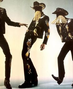 three men dressed in black and gold with long blonde hair, one wearing a cowboy hat