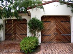 two wooden garage doors on the side of a white house with brick walkway and trees