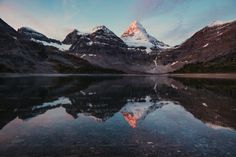 a mountain is reflected in the still water of a lake at sunset, with snow capped mountains in the background