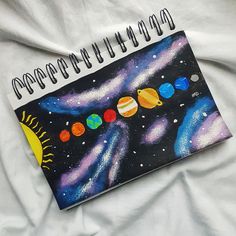 a spiral notebook with an image of the solar system painted on it, sitting on top of a white sheet