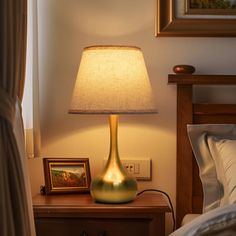 a lamp on a night stand next to a bed with a painting and framed pictures