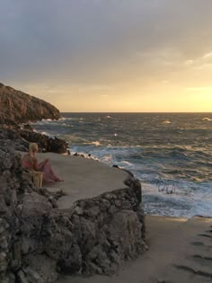 two people sitting on the edge of a cliff by the ocean at sunset or sunrise