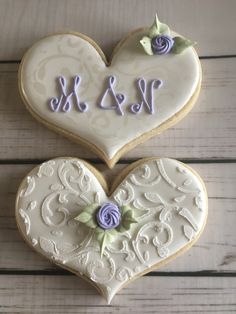 two decorated cookies in the shape of hearts with initials and flowers on them, sitting on a wooden surface