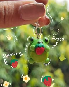 a hand holding a green frog keychain in front of some trees and flowers