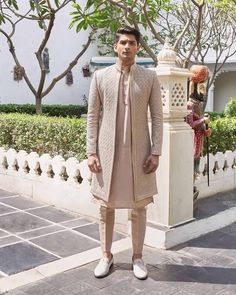 a man standing in front of a white fence wearing a tan outfit and shoes with his hands on his hips