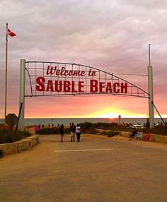 people walking under a welcome to sauble beach sign in front of the ocean