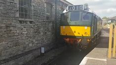 a green and yellow train traveling down tracks next to a brick building with two windows