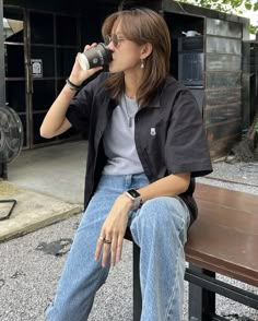 a woman is sitting on a bench drinking from a cup while wearing jeans and a black jacket