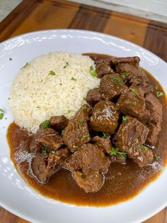 a white plate topped with meat and rice on top of a wooden table next to a knife