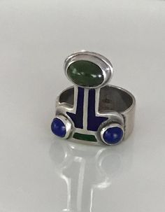 There is a hint of arts and crafts, as well as, modernist in this unusual ring which you can wear with the Peridot facing up or down! The beautiful green stone, as well as the smaller vibrant blue Lapis are all cabochon and bezel set. The enamel is in wonderful condition. Aside from some slight wear, the ring is in overall fabulous shape. The band is almost 1/4" wide, it is marked Sterling and has the initials CHM scratched on the silver behind the Peridot. Please see all photos for details, as Modernist Rings With Inlay For Gift, Modernist Rings With Inlay Perfect For Gifts, Art Deco Green Emerald Multi-stone Ring, Unique Enamel Rings With Gemstone, Modern Green Cabochon Jewelry, Unique Green Inlay Rings, Modern Green Multi-stone Jewelry, Modernist Multi-stone Ring, Modern Green Multi-stone Rings