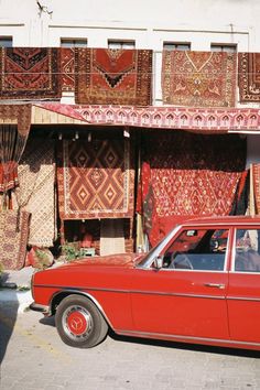 an old red car is parked in front of a building with many rugs on it