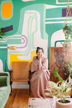 a woman sitting on a couch in front of a wall with colorful paintings and plants