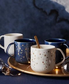 three blue and white coffee mugs on a gold tray with christmas lights around them