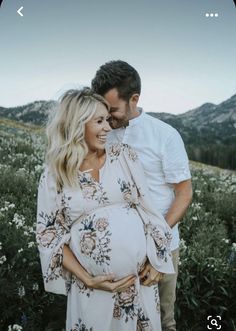 a pregnant couple standing in a field with mountains in the backgrouund and flowers all around them