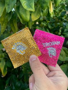 There is a green leafy background. In front of this background is a hand holding two Trojan condoms. The left one is gold colored and the right one is pink. These two items are bedazzled with rhinestones of their respective colors. Friends Laughing, Gag Gifts, Make Your Day, Get Started, Make Your, Make It Yourself, Art