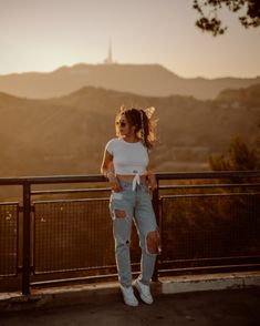 a woman standing on top of a hill next to a metal fence with mountains in the background