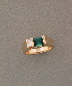 a gold ring with an emerald colored stone and two diamonds on the side, sitting on a gray surface