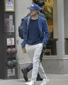 a man walking down the street wearing a blue jacket and white pants with a baseball cap on