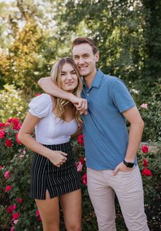 a man and woman standing next to each other in front of pink flowers with their arms around each other