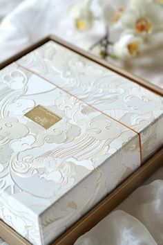 an open white box with gold trim sitting on top of a bed next to flowers