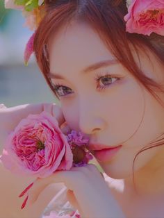 a woman with flowers in her hair holding a pink flower up to her face and looking at the camera