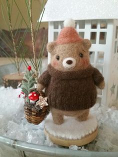 a teddy bear with a knitted hat and sweater is sitting in the snow next to a small christmas tree