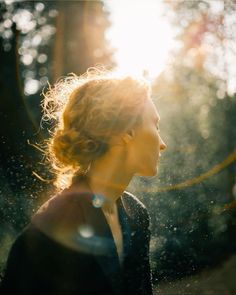 a woman standing in front of trees with the sun shining down on her face and hair blowing in the wind