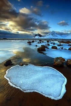 an icy lake with rocks and snow on the ground in front of a snowy mountain