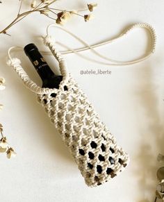 a white crocheted bag with a black bottle in it next to some flowers