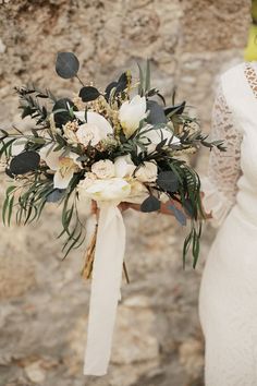 a bride holding a bridal bouquet with greenery and white flowers in front of a stone wall