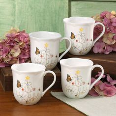 three white coffee mugs with butterflies on them sitting next to some pink and yellow flowers