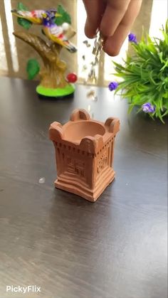 a hand is reaching for a small clay pot on a table with flowers in the background