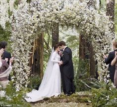 a bride and groom standing under an arch with white flowers on it in the woods