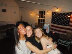 three girls hugging each other in a room with an american flag hanging on the wall