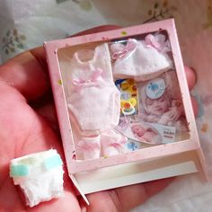 a hand holding a small doll in a pink box with its clothes and accessories inside