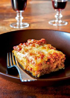 a piece of lasagna on a plate with a fork and two glasses of wine in the background