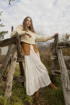 a woman leaning on a wooden fence wearing a white dress and brown boots with her arms crossed